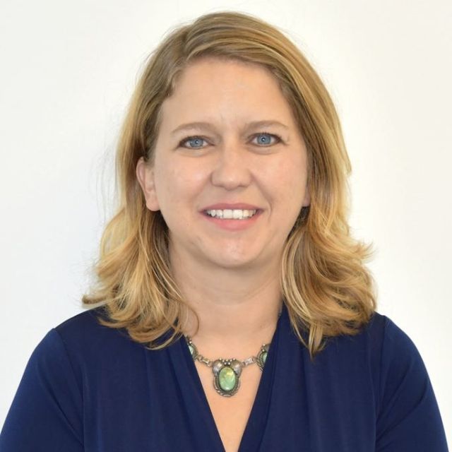 Portrait of a woman with shoulder-length blonde hair, wearing a navy blue top and a green stone necklace, smiling against a plain white background.
