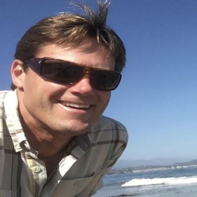 The image shows a man outdoors near the ocean on a sunny day. He is smiling, wearing sunglasses and a plaid shirt, with blue sky and waves visible in the background.